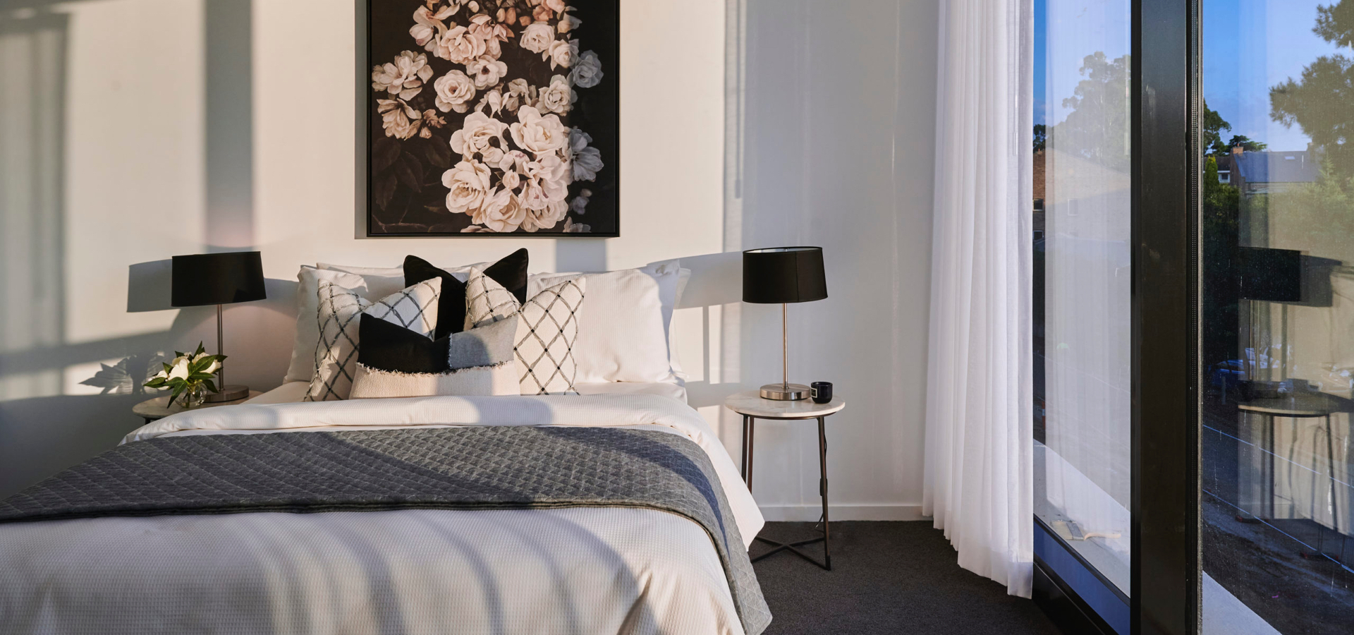 A bed with a black and white picture above it and table and lamp to the right of it.