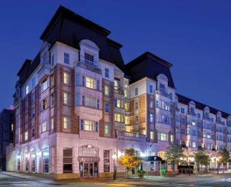Front view of a five-story, tan and white, hotel at dusk with the name “Hotel Commonwealth” and “Island Creek Oyster Bar” above the front doors. 