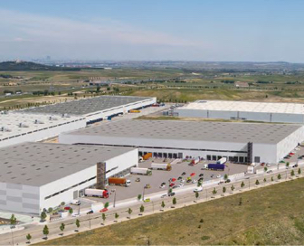Aerial view of a large grey and white logistics/industrial building with a parking lot with trucks and a road in front of it. 