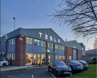 A small, red brick and grey tin logistics building with the name “Tesla” above the front door with a parking lot of cars in front of it.
