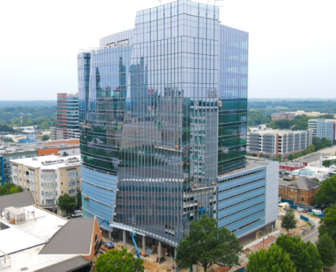 A large, blue, glass office building with trees in front of it and other small city buildings surrounding it.
