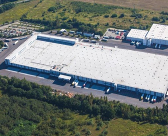 Aerial view of a large white logistics building with trees, a full parking lot and truck bays surrounding it.